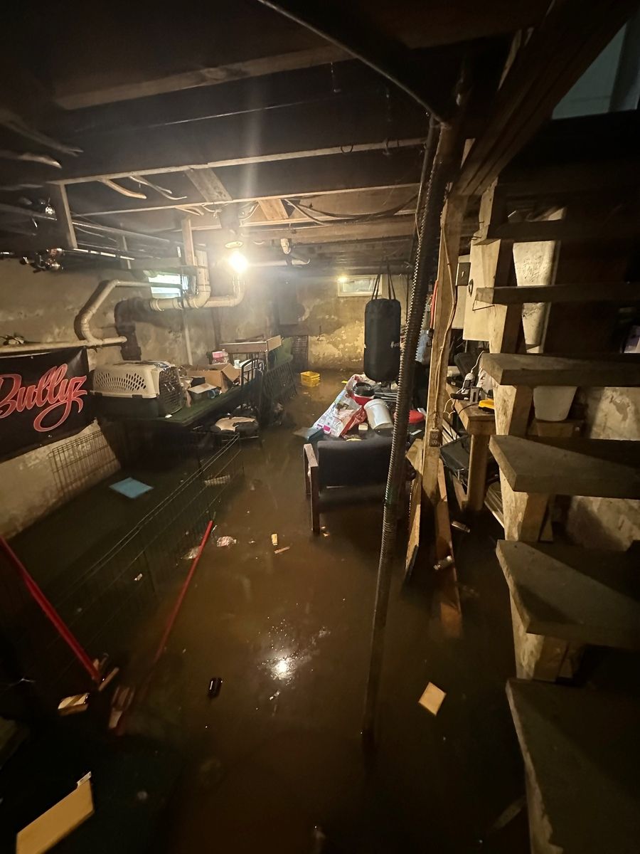 Flooded basement with debris. Water covers the floor, with a staircase and cluttered furniture. Dark, dim lighting.