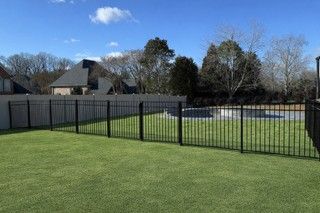 Black metal fence encloses a green lawn with a pool, houses, and trees in the background under a blue sky.