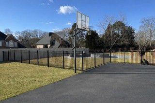 Black wrought-iron fence surrounds asphalt basketball court and grassy yard on a sunny day.