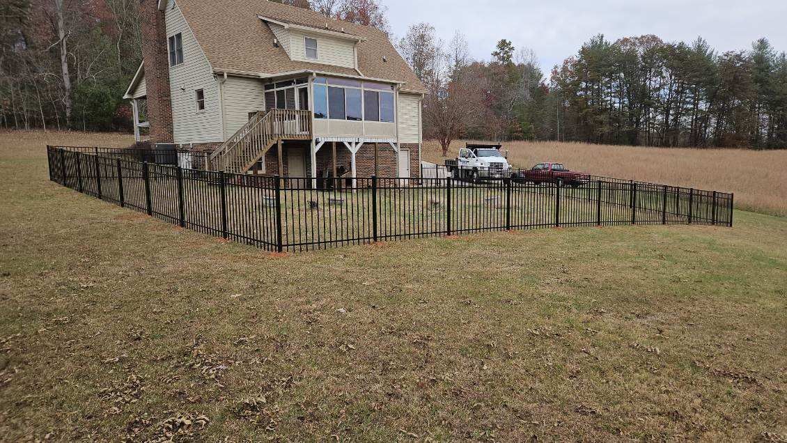 Beige house with porch and wooden fence in a field; a car is parked nearby.