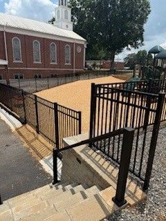 Exterior view of a church with a fenced playground and steps. Dark metal fence and beige gravel.