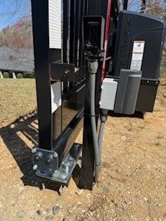 Black gate post with attached control box, electrical conduit, and security camera, set in dirt.