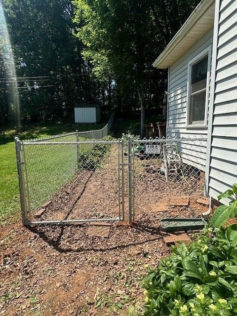 A chain-link fence encloses a dirt area next to a house with light blue siding.