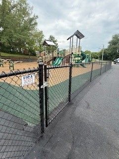 A fenced playground with slides and climbing structures. Gate is locked. Green and brown surfaces. Overcast sky.