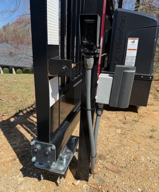 Black metal gate with mounted camera, control box, and gate operator in an outdoor setting.