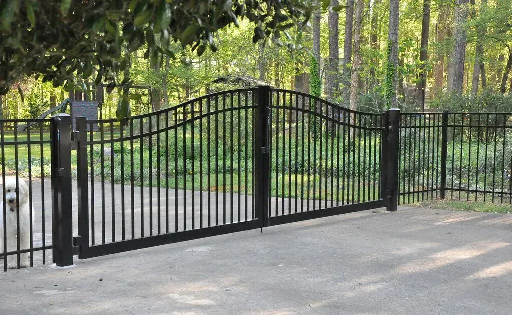 Black metal driveway gate with arched top and vertical bars, set against a blurred background of trees.