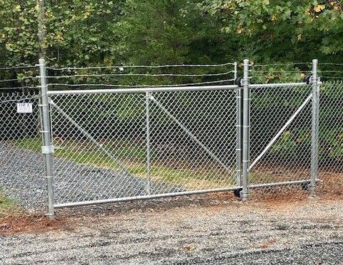 Chain-link gate with barbed wire on top, set in gravel driveway with wooded background.
