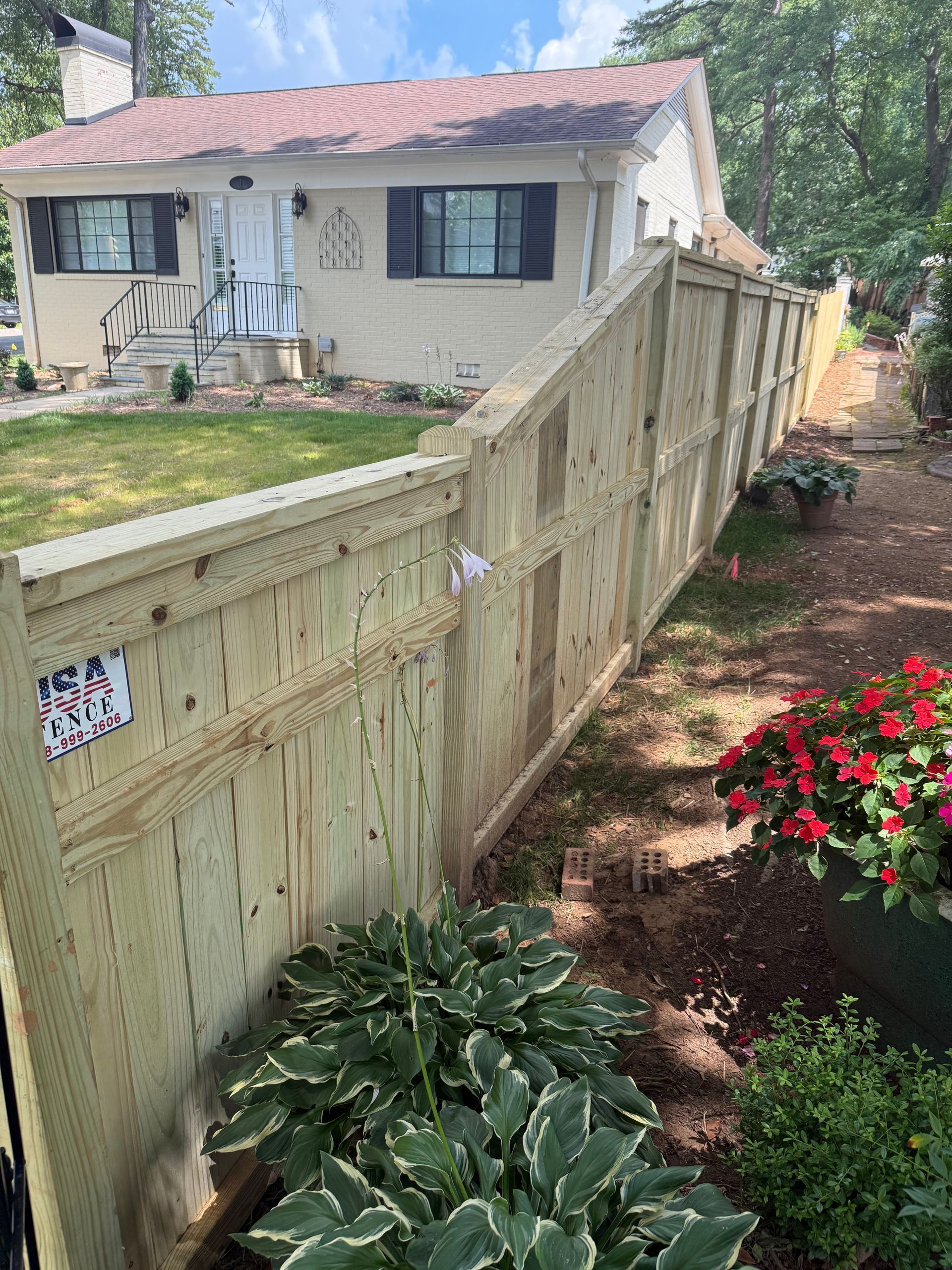 A wooden fence beside a light-colored house with green lawn and flowerbeds under a blue sky.