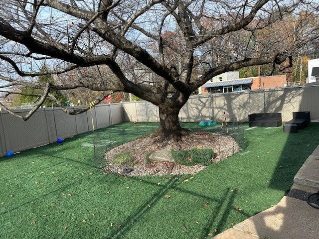 A backyard with artificial green turf, a large tree, and a stone-covered base. Grey fence surrounds.