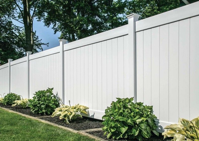 White vinyl fence with green bushes in front, against a backdrop of trees.