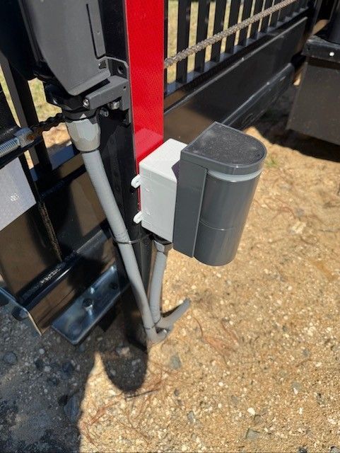 Sliding gate with a mounted photoelectric sensor and wiring. Black, gray, red, and white components on a gravel surface.