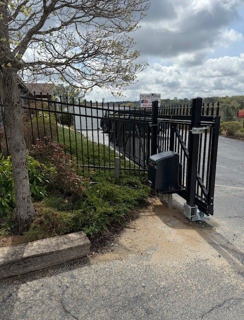 Black metal fence with an open automatic gate beside a tree and small bushes on a paved surface.