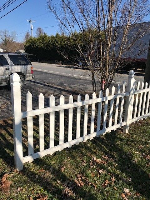 White picket fence in front of a road, with a tree and a car.