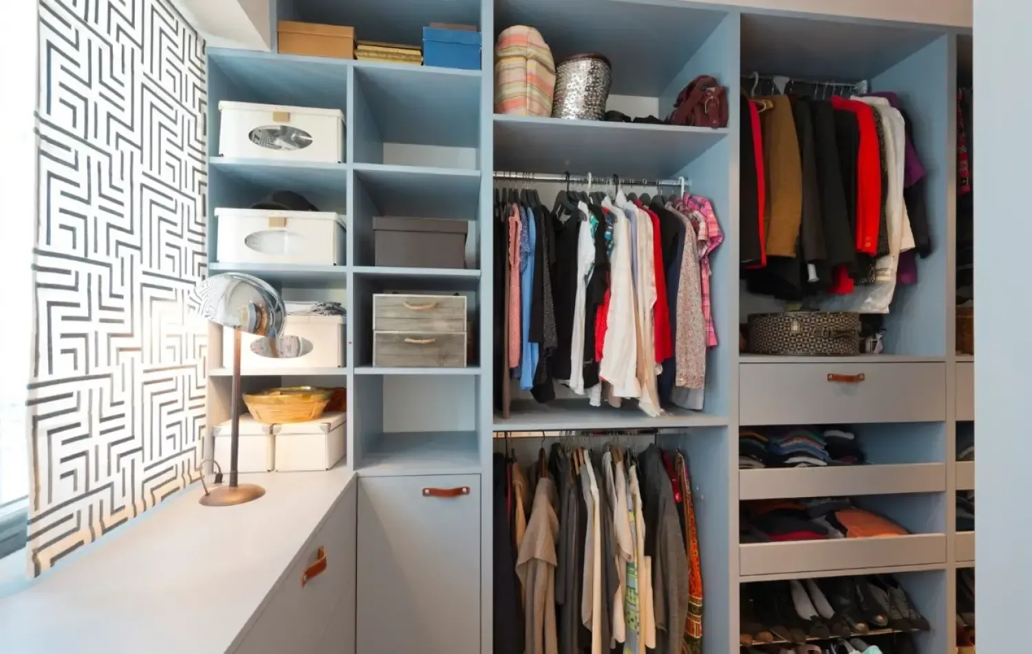 Blue built-in closet with shelves and hanging rods, filled with clothes and storage boxes, against a patterned wall.