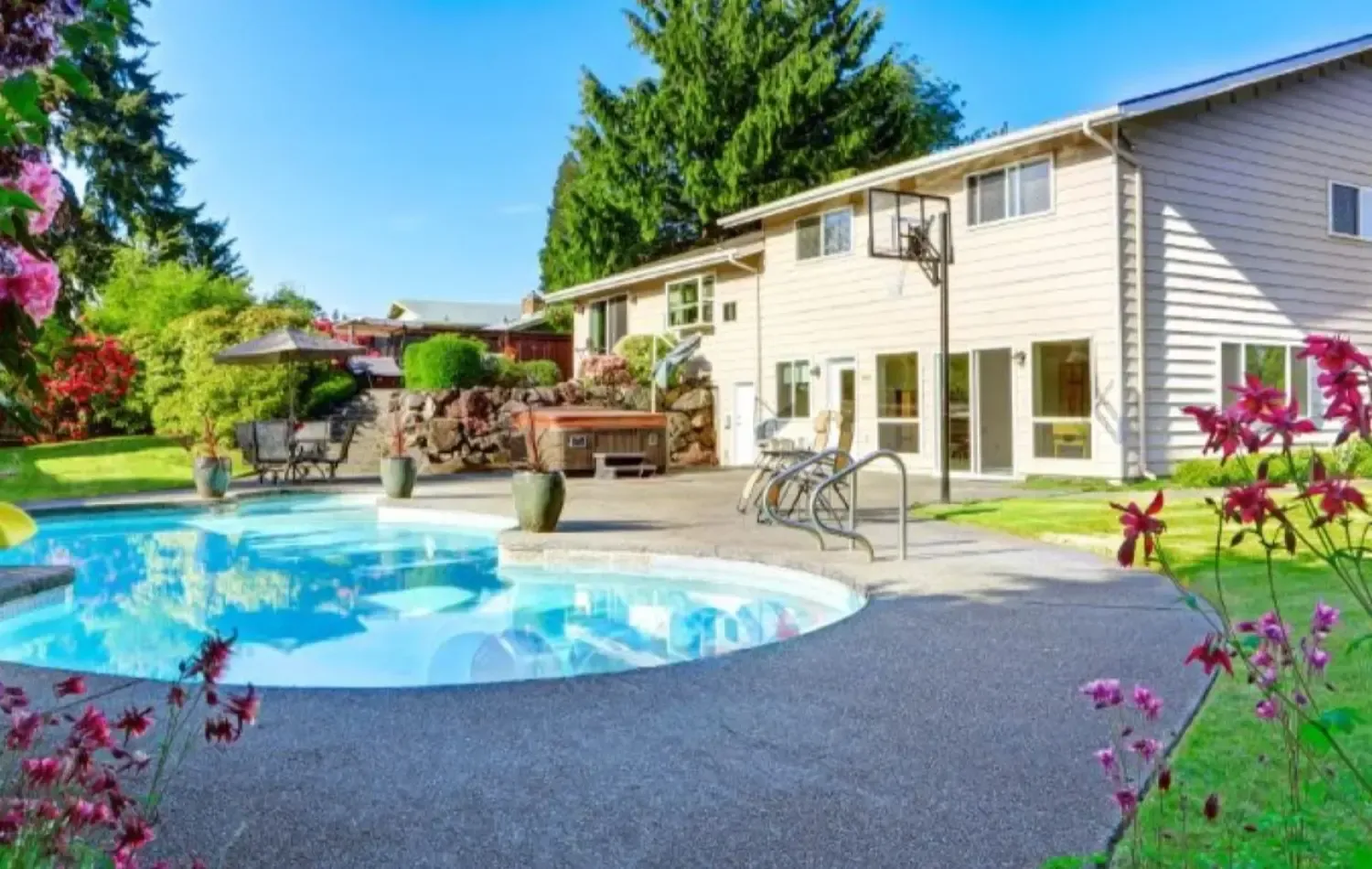 Backyard with a swimming pool, patio, hot tub, and two-story house with green landscaping.