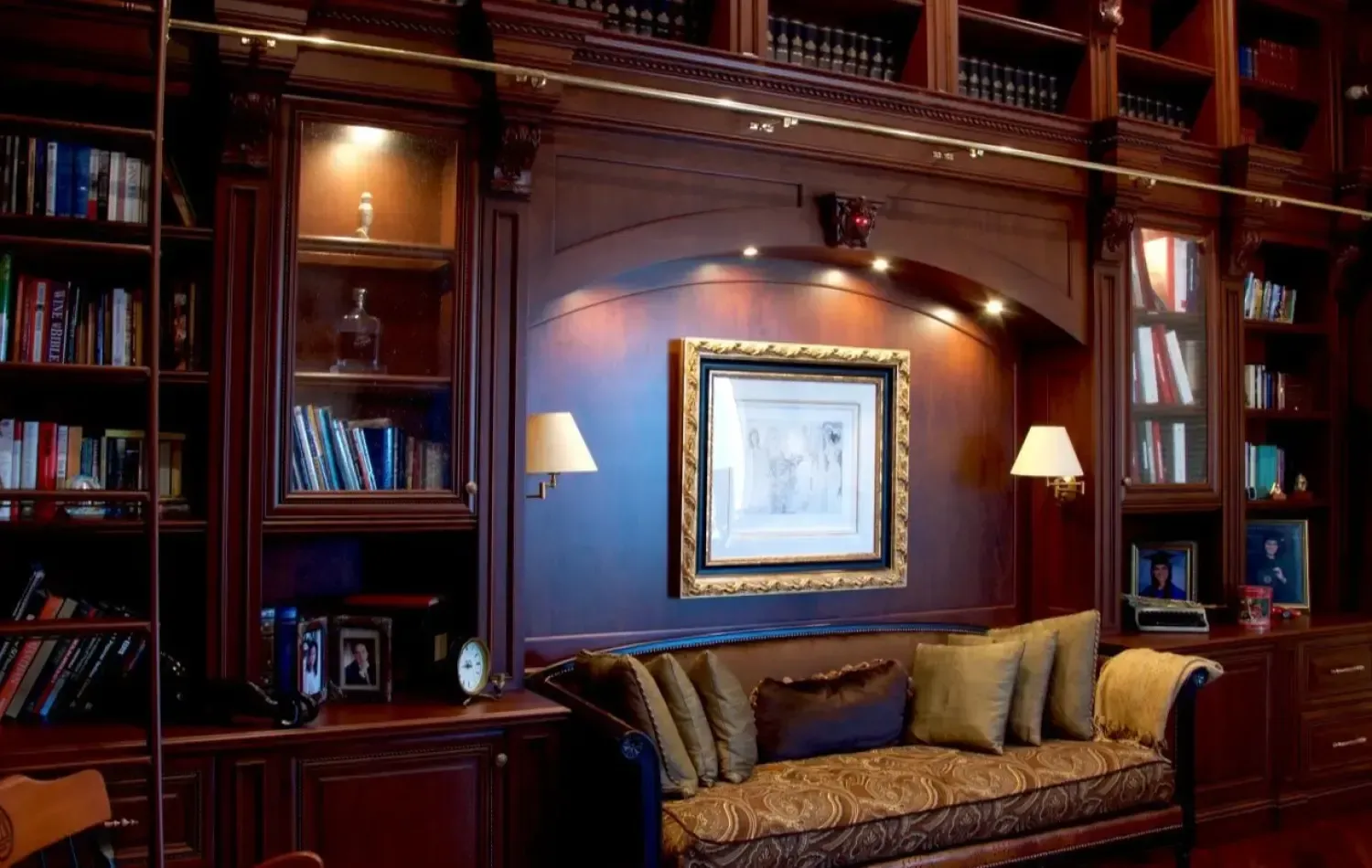 Dark wood library with built-in bookshelves, couch, and framed artwork. Warm lighting accents the space.