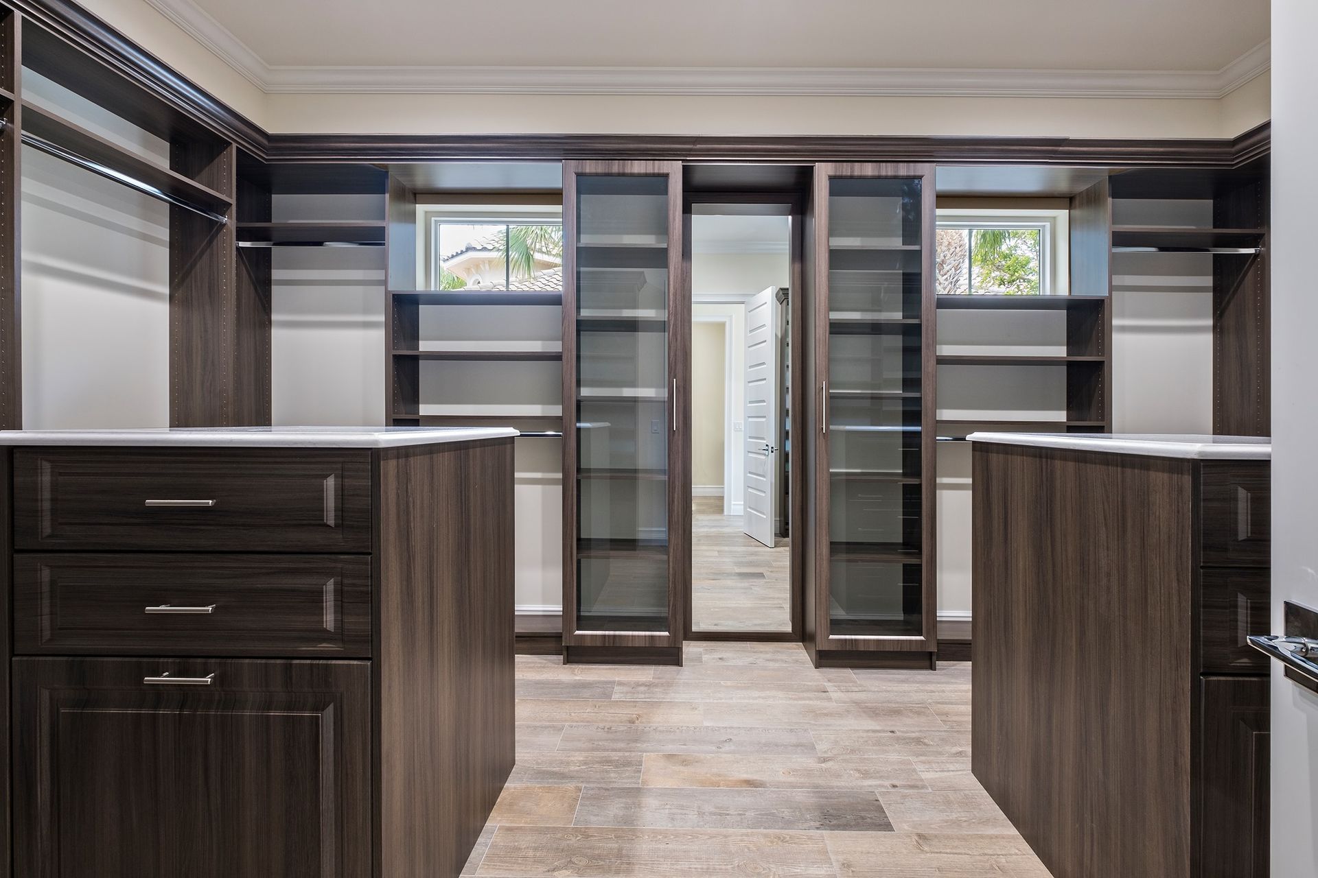 Walk-in closet with dark wood cabinetry, glass-door sections, and islands. Light stone flooring and white accents.