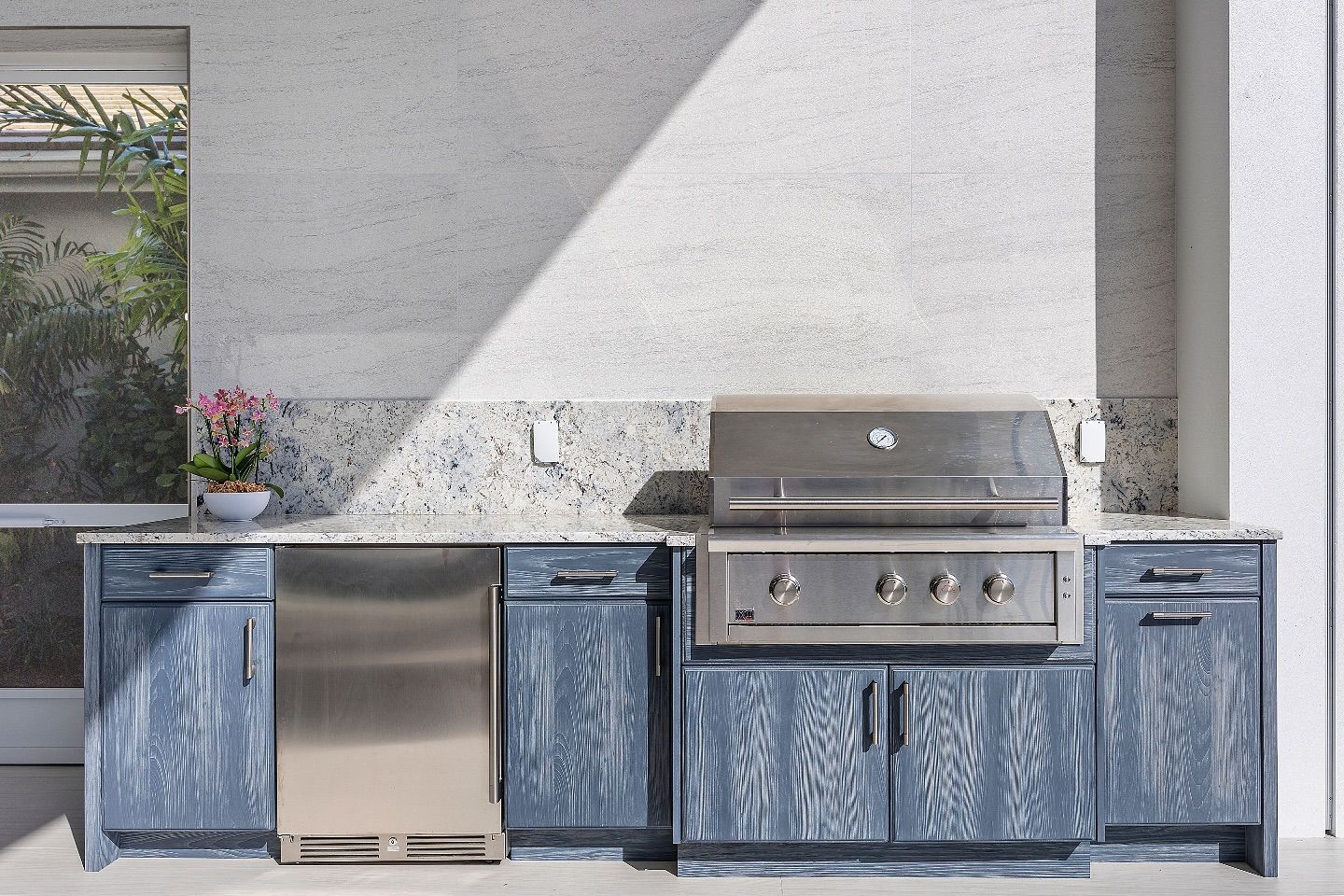 Outdoor kitchen with blue cabinets, stainless steel grill, and refrigerator against speckled wall.