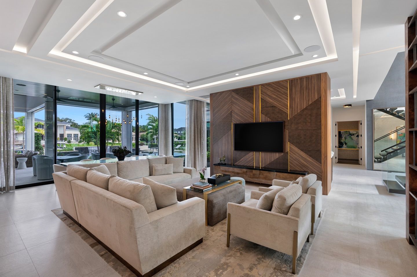 Modern living room with beige sofas, a large TV, and floor-to-ceiling windows overlooking a green landscape.