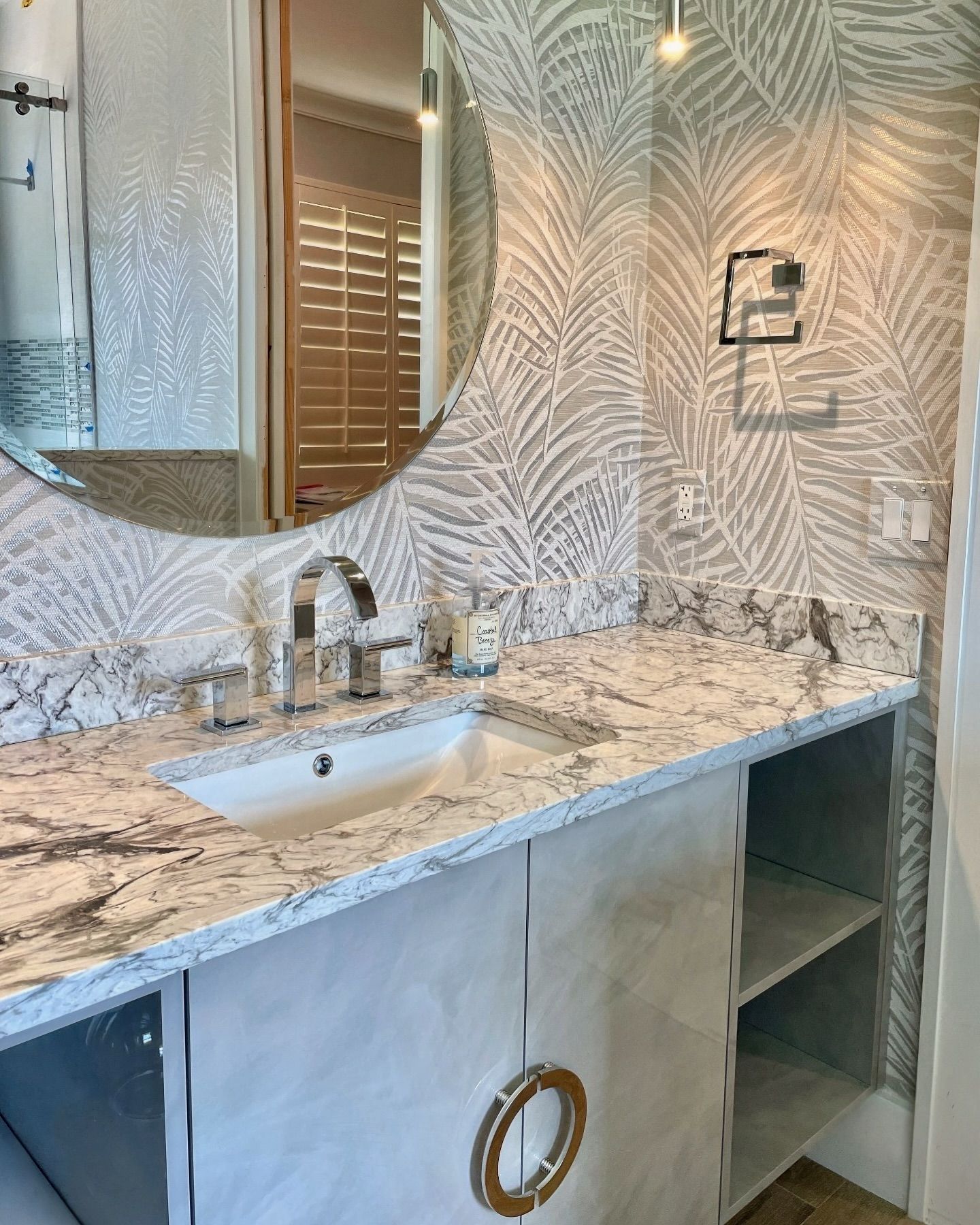 Bathroom with marble countertop, sink, round mirror, and palm leaf wallpaper.