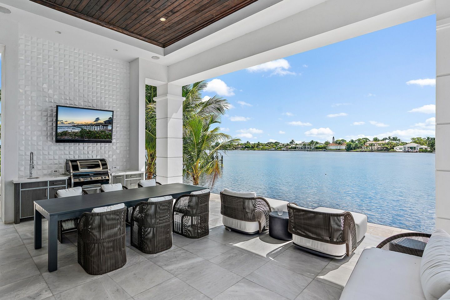 Patio overlooking water, with a dining table, chairs, and a TV; a serene, sunny day.