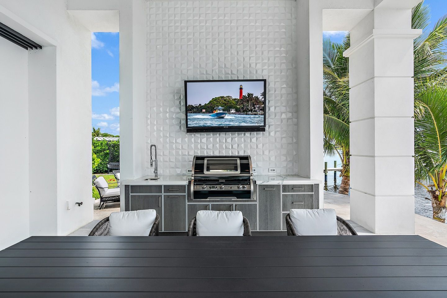 Outdoor kitchen with grill, sink, and TV; white tile backsplash; view of water and trees.