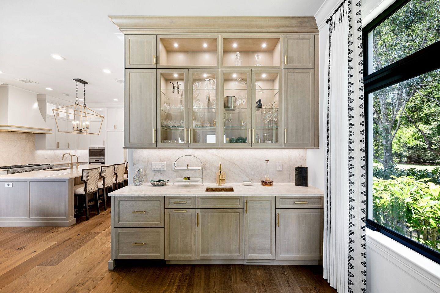 Luxury home bar with glass-front cabinets, marble countertop, and gold fixtures. Window with view to lush green outdoors.