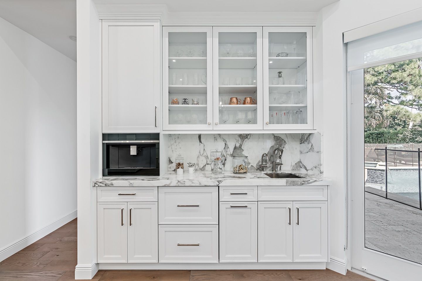 White built-in coffee/bar station with glass-front cabinets, marble backsplash and countertop, and a window overlooking a pool.