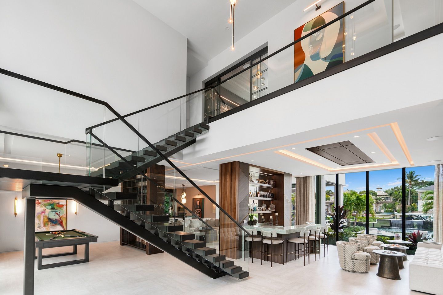Modern, two-story interior with black staircase, glass railings, bar area, and large windows overlooking water view.