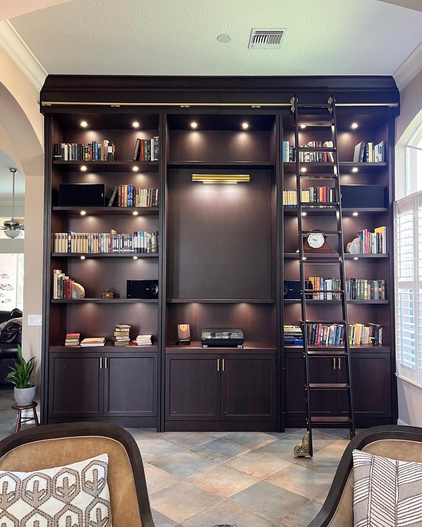 Dark wood built-in bookcase with ladder, books, and lighting. Dark brown cabinets with gold accents are set in a home.