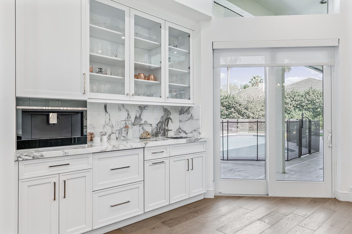White kitchen cabinets with glass doors, marble backsplash, built-in coffee machine, and view of a pool.