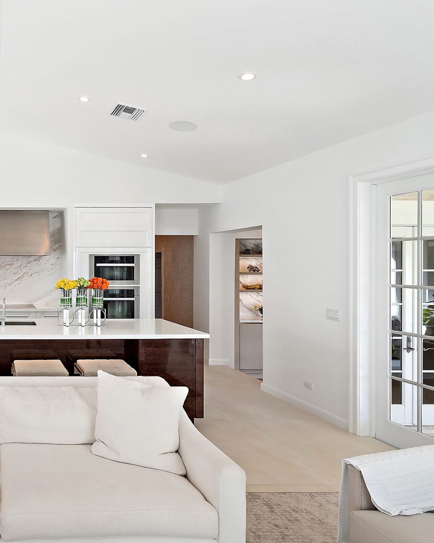 Spacious modern kitchen with white walls, island, and seating. Light wood floors, sofa, and French doors.