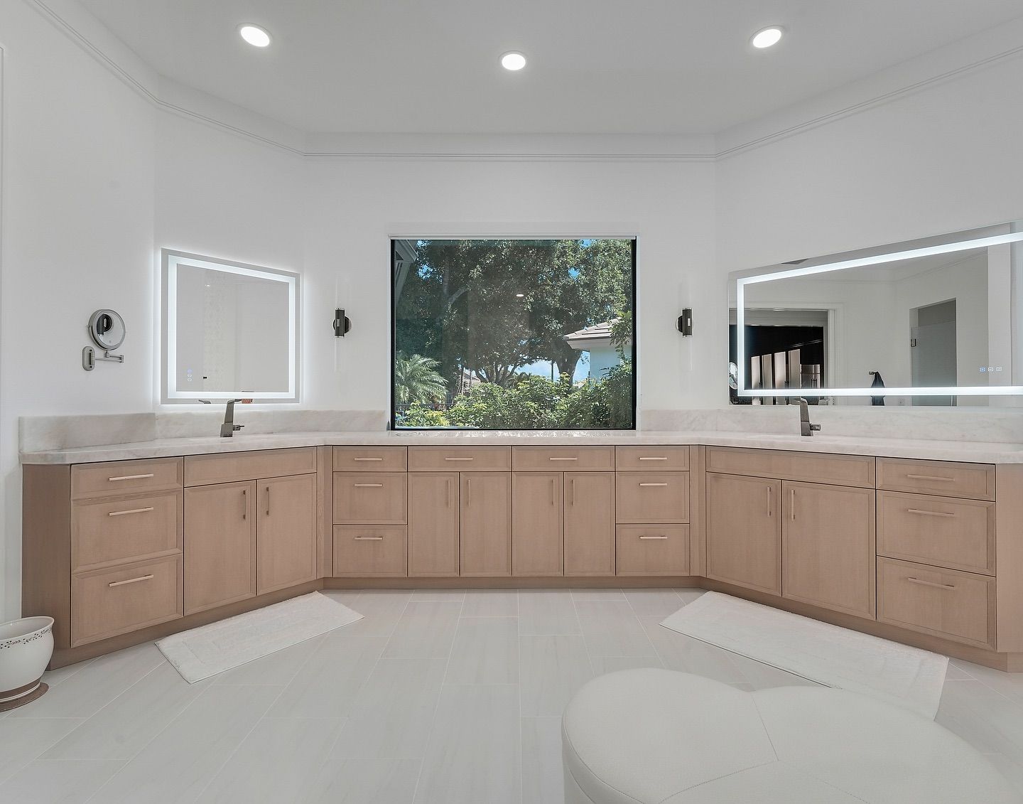 Spacious white bathroom with light wood cabinets, large window, and two illuminated mirrors.