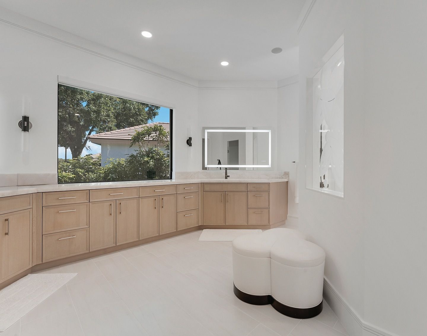 Bathroom with large window, light wood cabinets, white walls, and a stool.