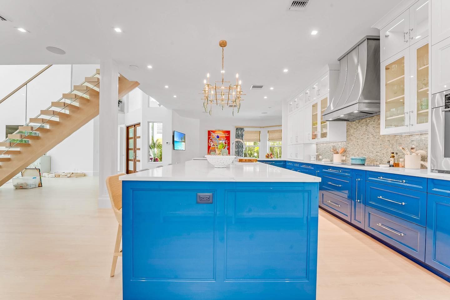 Bright blue kitchen with white countertops and island, chandelier, light wood floors, and staircase.