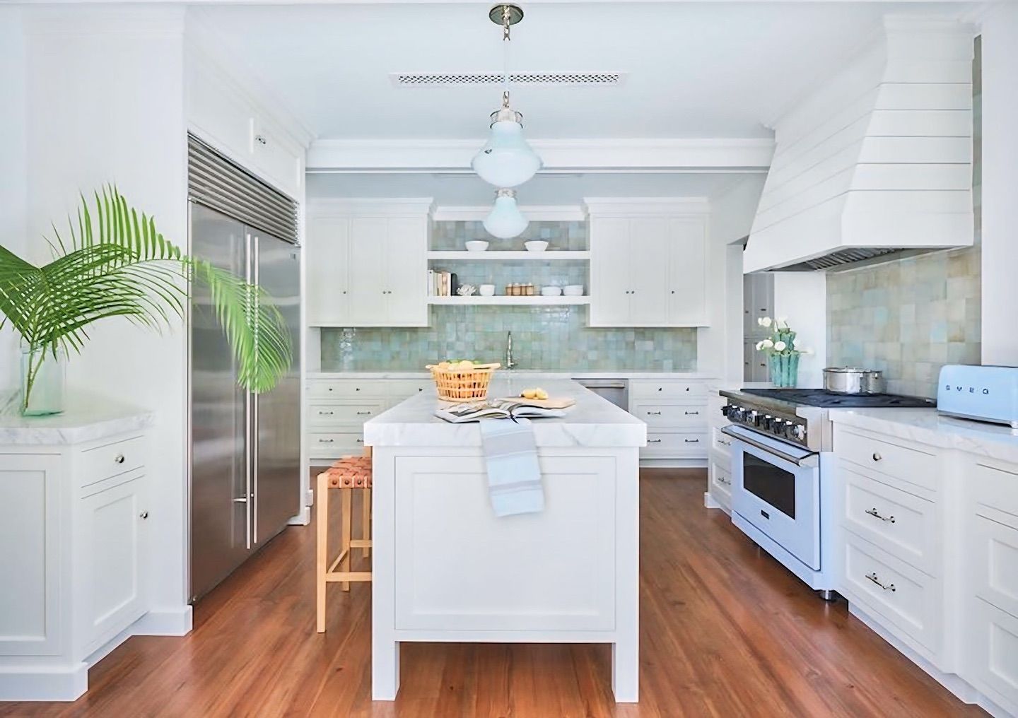 Bright white kitchen with a marble-topped island, stainless steel refrigerator, and blue tiled backsplash.