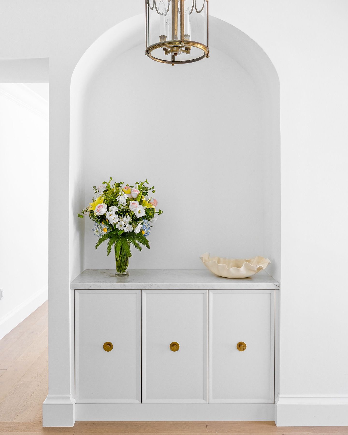 White built-in cabinet with marble top, arched alcove, floral arrangement, shell, and brass light fixture.
