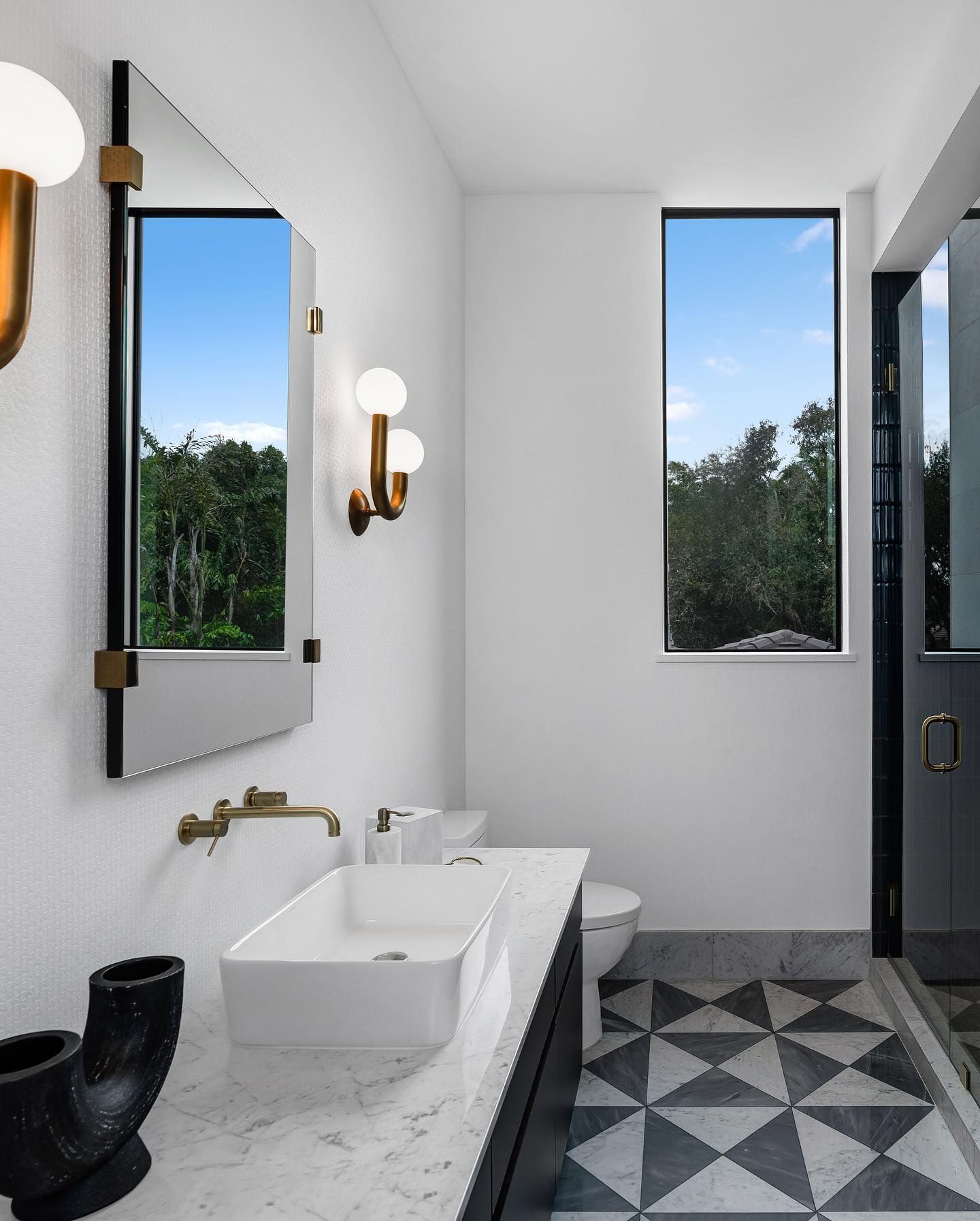 Modern bathroom with white walls, geometric tile floor, large mirror, and sink, with a window offering a view of trees.