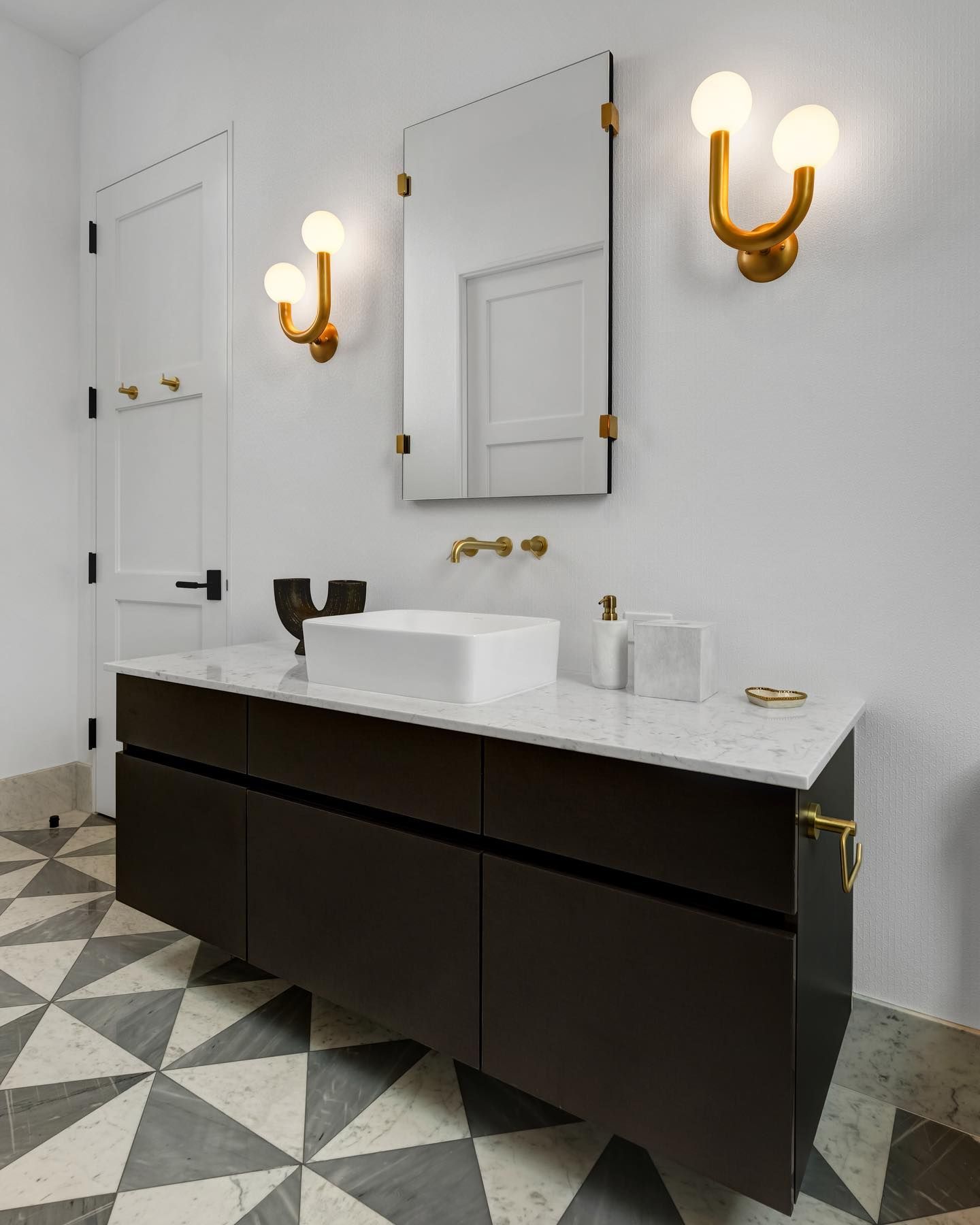 Bathroom with modern design: dark vanity, white countertop, rectangular mirror, gold sconces, patterned floor.