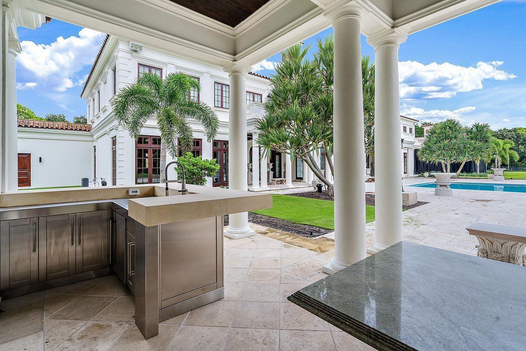 Outdoor kitchen with stainless steel appliances under a covered patio, overlooking a mansion and pool.