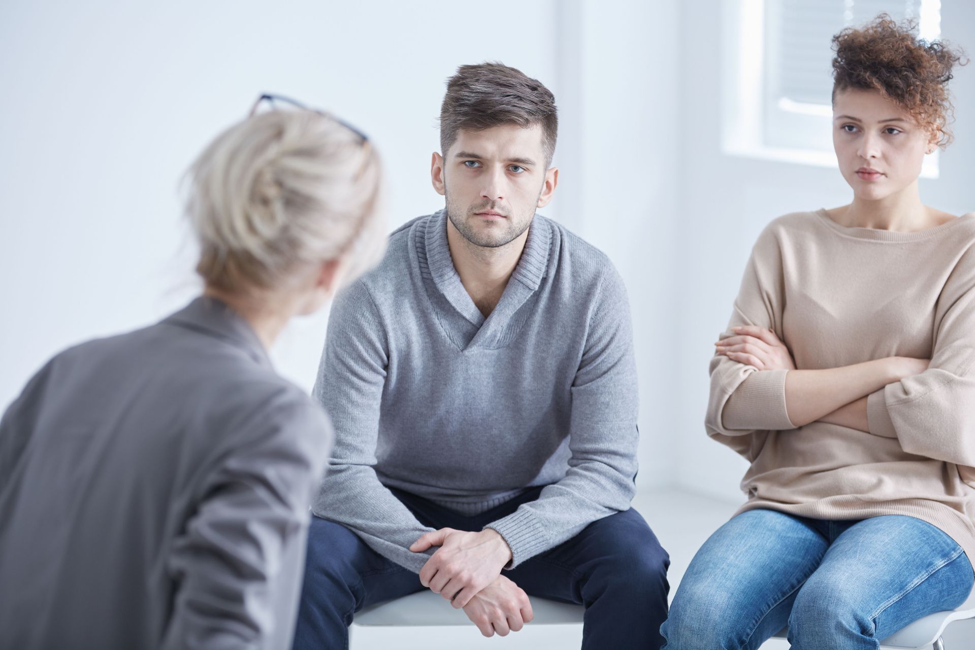 Two people sit across from a counselor in a bright office, looking tense and attentive.