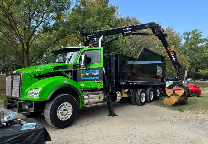 Green and black tree service truck with crane lifting a tree trunk.