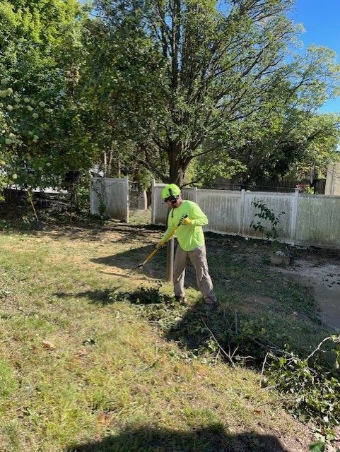Person raking a yard, wearing a lime green shirt and hat. Tall tree and white fence in background.