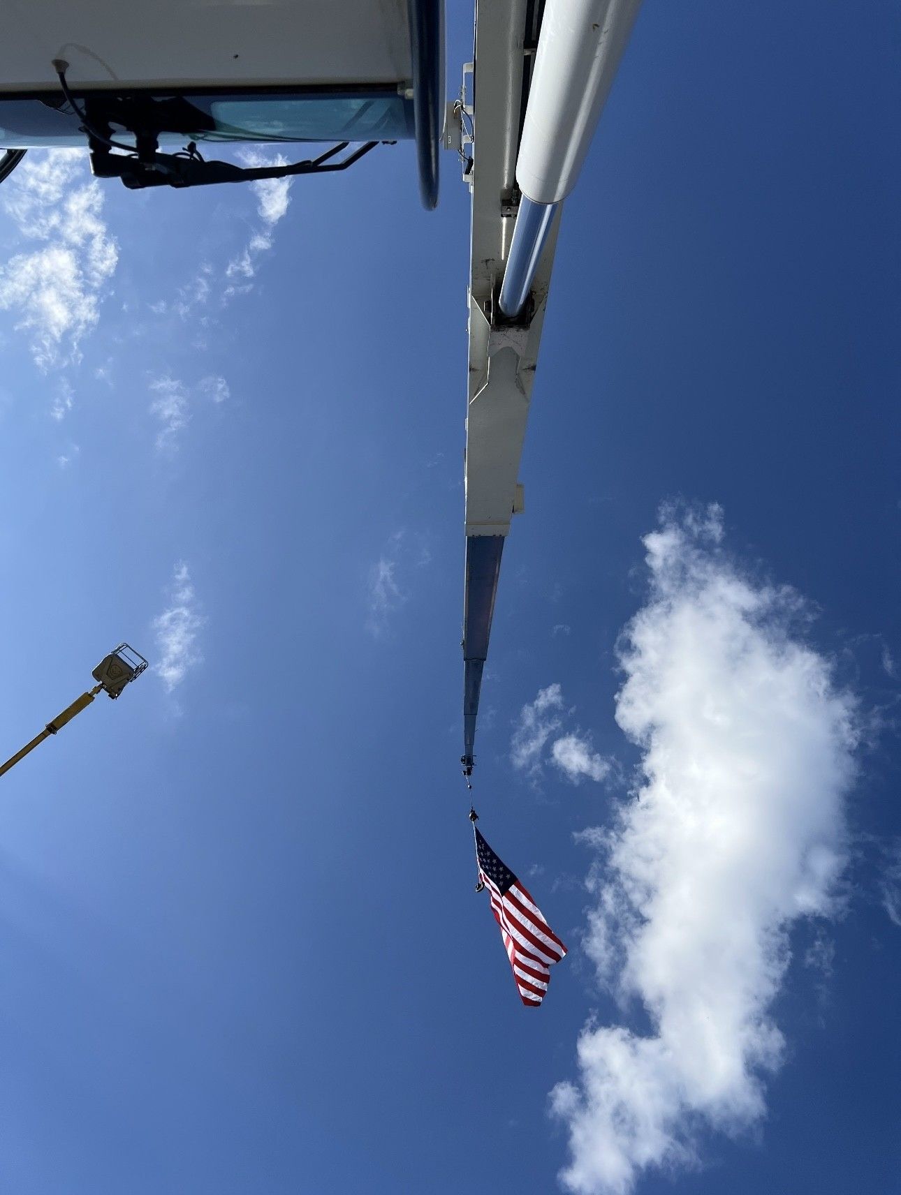 American flag hanging from a tall blue crane against a bright blue sky with clouds.