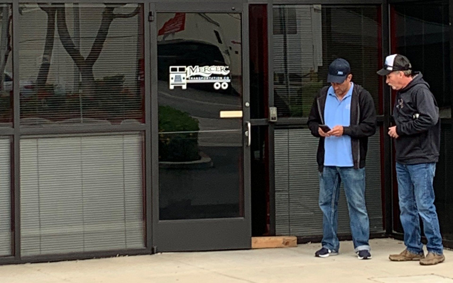 Two men stand outside a storefront, one looking at his phone while the other talks beside him.