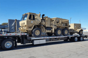 Tan military vehicle loaded on a flatbed trailer in a parking lot under a clear blue sky