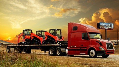 Red semi truck hauling two orange excavators on a flatbed at sunset
