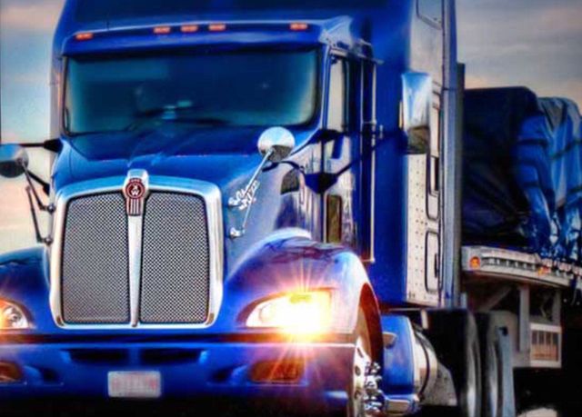 Blue semi-truck with headlights on, towing a covered trailer on the road