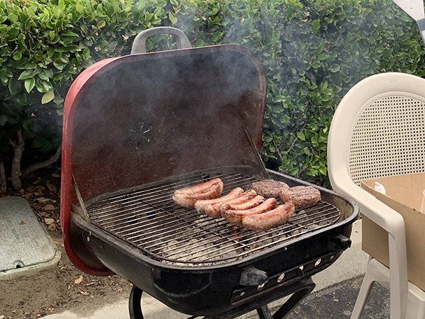 Outdoor charcoal grill with sausages cooking beside a beige chair, smoke rising in a backyard.