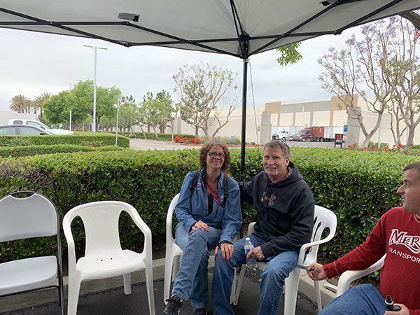 Three people sitting under a canopy outdoors, with chairs and hedges in the background.